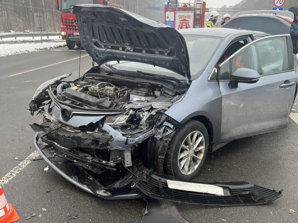 Rozbity samochód osobowy z otwartą maską stoi na drodze, obok widać wozy strażackie oraz grupę ratowników.