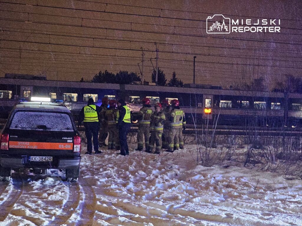 Grupa strażaków w uniformach omawia sytuację w pobliżu torów kolejowych, obok zaparkowanego pojazdu służb ratunkowych.