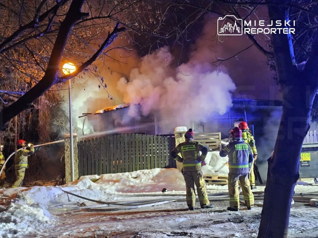 Grupa strażaków w hełmach gasi pożar budynku, z którego unosi się gęsty dym, w otoczeniu śniegu i latarni.