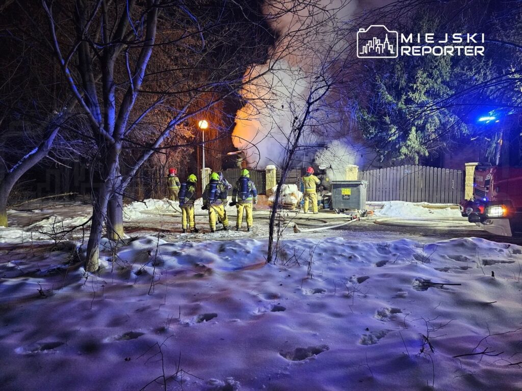 Grupa strażaków w żółtych kombinezonach gasi pożar w nocy, otoczona ośnieżonym terenem i drzewami.