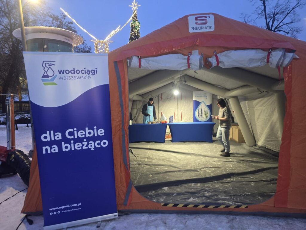 Pracownicy w namiocie informacyjnym przygotowujący stoisko promocyjne, z banerem "Wodociągi Warszawskie" w tle.