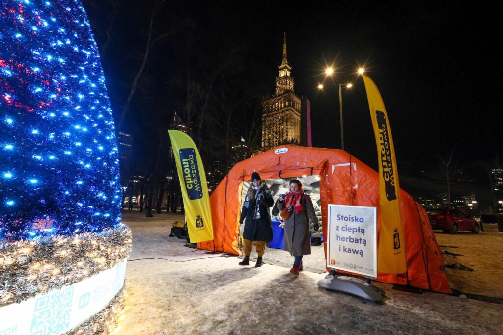 Kobieta w płaszczu i mężczyzna w ciepłej odzieży wychodzą z pomarańczowego namiotu z napisem „Stoisko z ciepłą herbatą i kawą” przy rozświetlonej choince.