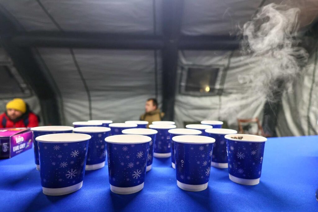 Na stole znajdują się niebieskie kubki papierowe z białymi śnieżynkami, a w tle widać zamgloną postać siedzącą w namiocie.