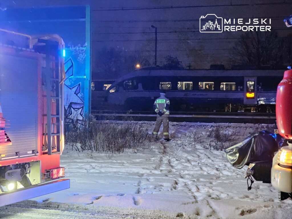 Strażak w odblaskowym uniformie obserwuje torowisko w nocy, obok stoi wóz strażacki z włączonymi sygnałami.