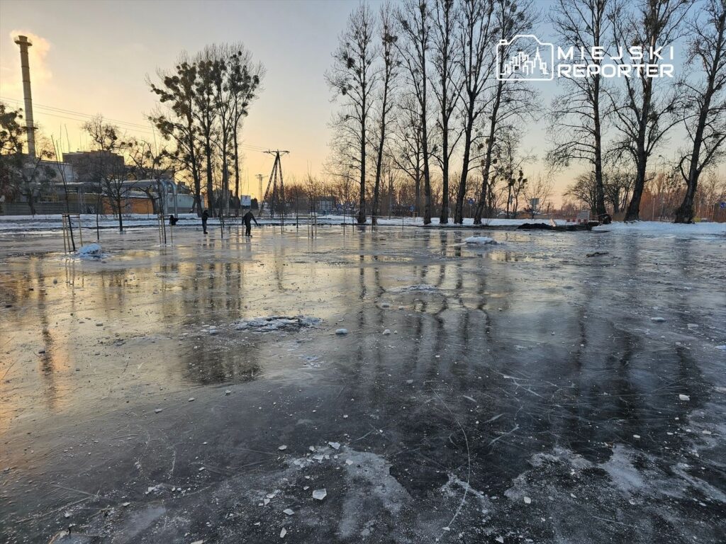 Osoby poruszające się po zamarzniętej tafli lodu w parku, otoczone drzewami i przemysłowym tłem.