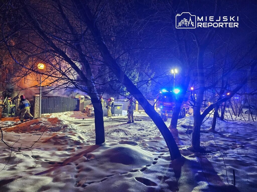 Strażacy gaszą pożar w pobliżu ogrodzenia, otoczeni przez dym i śnieg, w nocy przy włączonych sygnałach świetlnych.