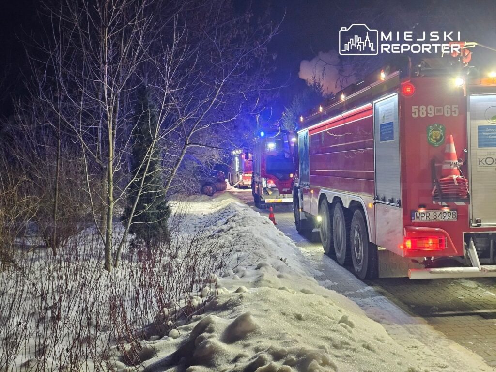 Strażacy w wozach gaśniczych pracują w zaśnieżonym terenie nocą, w tle widoczne dymiące miejsce.
