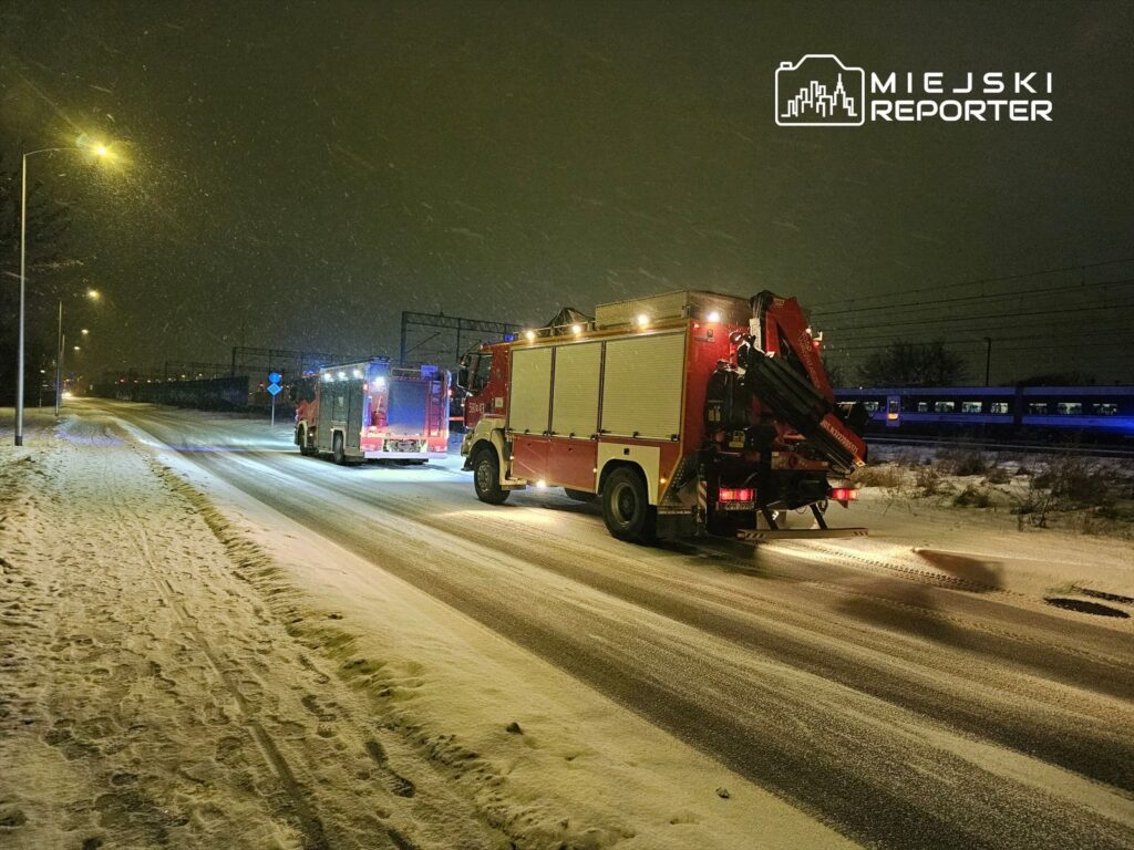 Czerwony wóz strażacki zaparkowany na ośnieżonej drodze w nocy, obok drugiego wozu, w tle pociąg.