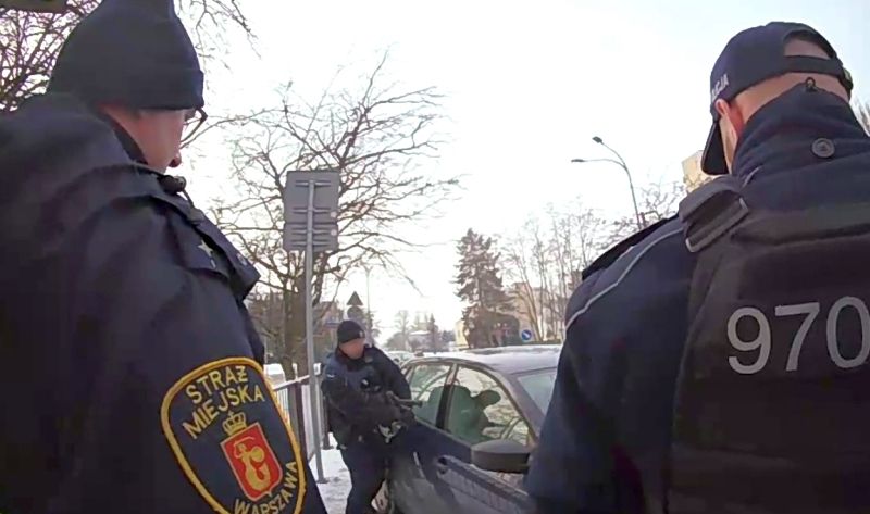 Dwaj funkcjonariusze straży miejskiej kontrolują zaparkowany samochód na ulicy w zimowej scenerii.