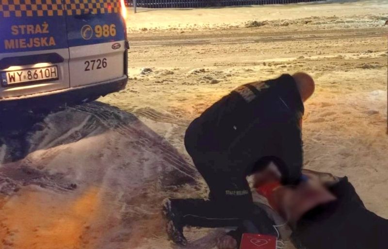 Funkcjonariusz straży miejskiej udziela pomocy osobie leżącej na zaśnieżonym chodniku obok radiowozu.