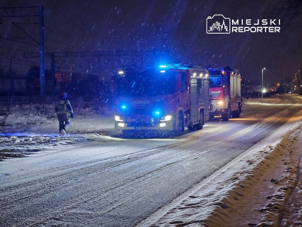 Strażacy przy wozie strażackim z niebieskimi światłami w śnieżnej scenerii nocnej na miejskiej drodze.