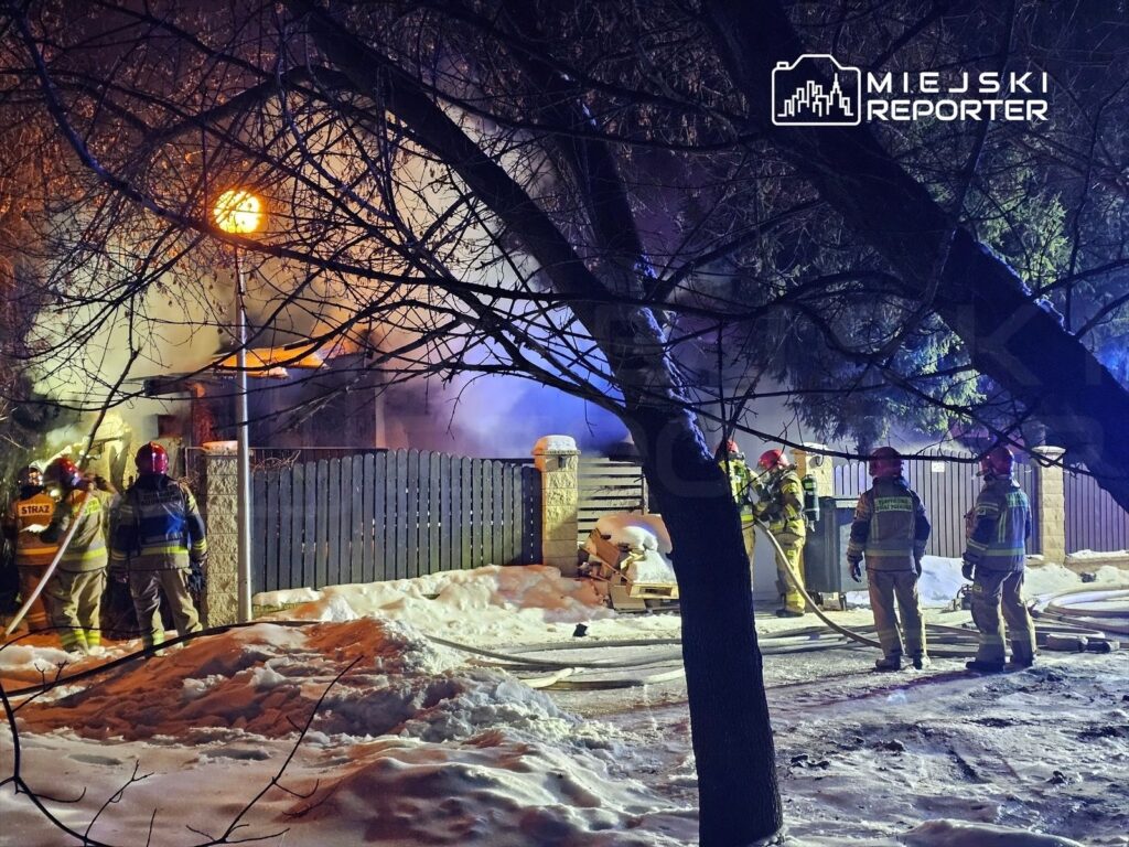 Grupa strażaków w hełmach walczy z pożarem budynku w zimowej scenerii, otoczeni przez dym i płomienie.