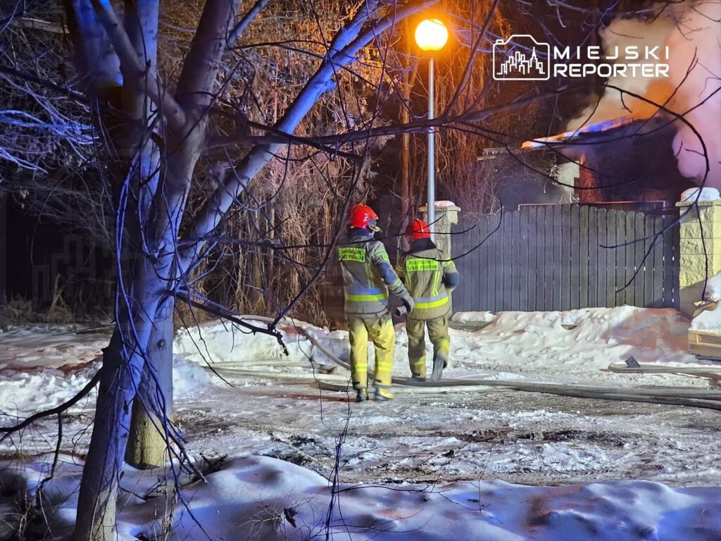 Dwaj strażacy w hełmach podchodzą do płonącego budynku w zimowym otoczeniu, oświetleni przez latarnię.