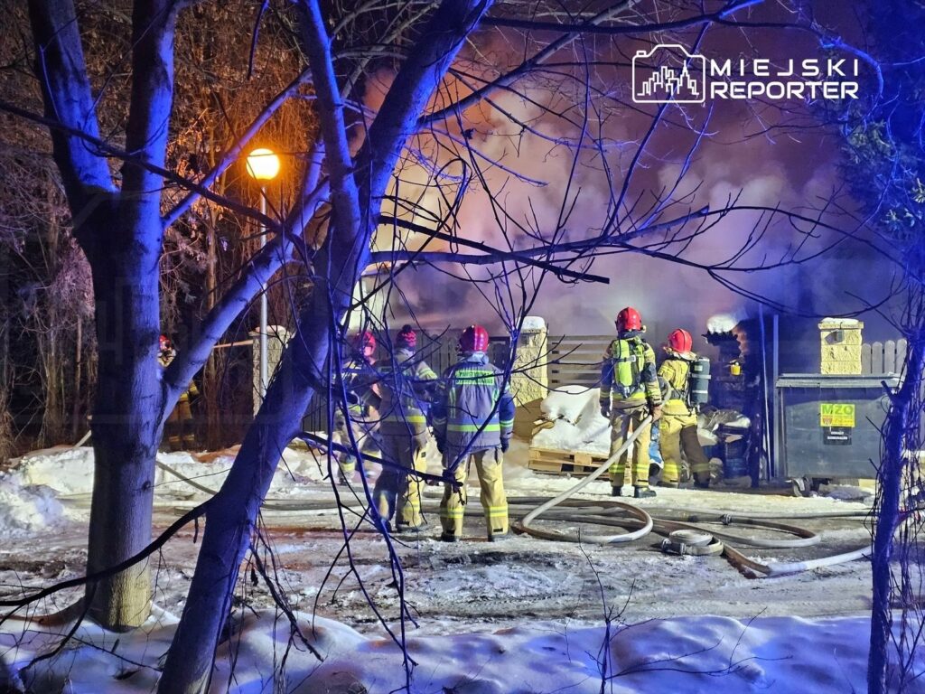 Grupa strażaków w hełmach gasi pożar w pobliżu kontenera na odpady, w tle widoczne unoszące się dym.