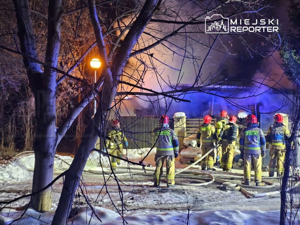Grupa strażaków w hełmach gasi płonący budynek w zimowej scenerii, otoczeni dymem i drzewami.