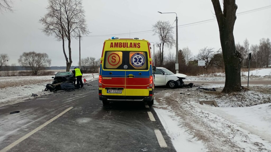Policyjny funkcjonariusz bada miejsce wypadku drogowego na zaśnieżonej drodze, obok uszkodzonych pojazdów i karetki.