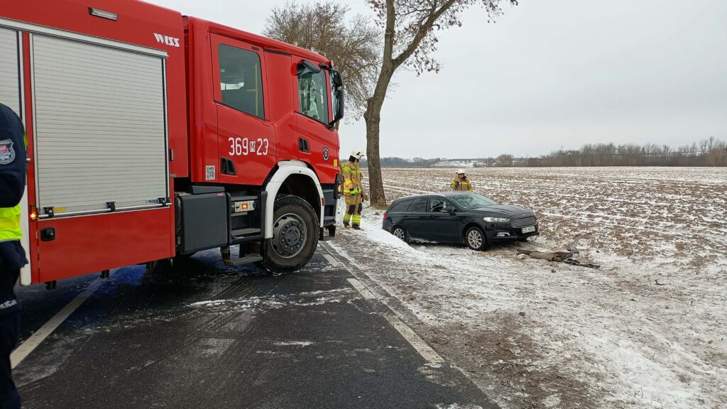 Czterech strażaków w mundurach ocenia sytuację przy czarnym samochodzie, który wpadł do rowu na zaśnieżonej drodze.