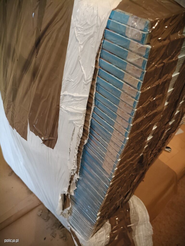 Uszkodzony karton z niebieskimi paczkami wypełnionymi przedmiotami, otoczony brązowym i białym materiałem.