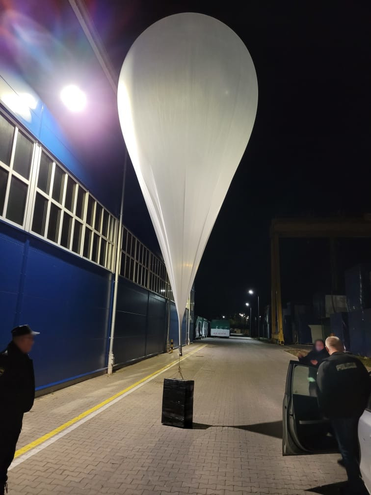 Biały balon meteorologiczny unosi się nad chodnikiem w nocy, obok dwóch mężczyzn w mundurach stojących przy samochodzie.