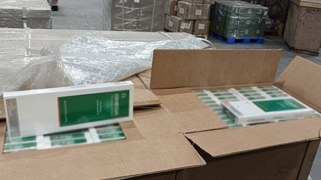 Otwarte kartony z produktami medycznymi na tle magazynu, w jednym z nich widoczny biały opakowanie z zielonym nadrukiem.