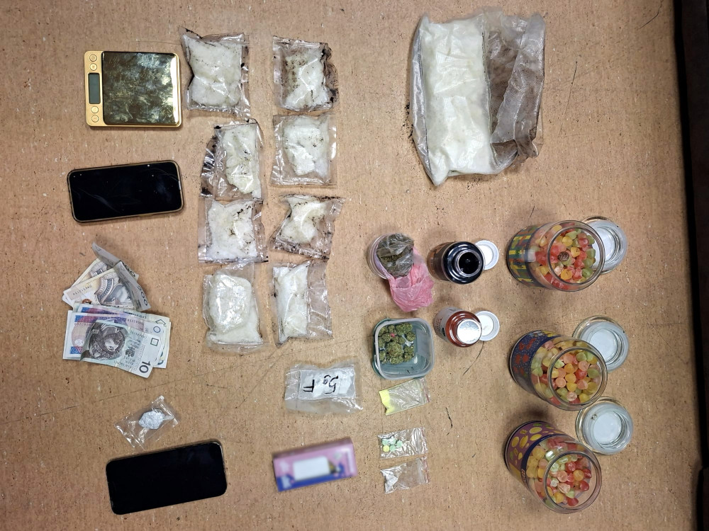 Na blacie leżą różne opakowania z substancjami, w tym białe proszki, słoiki z cukierkami oraz gotówka. Obok znajdują się dwa telefony komórkowe.