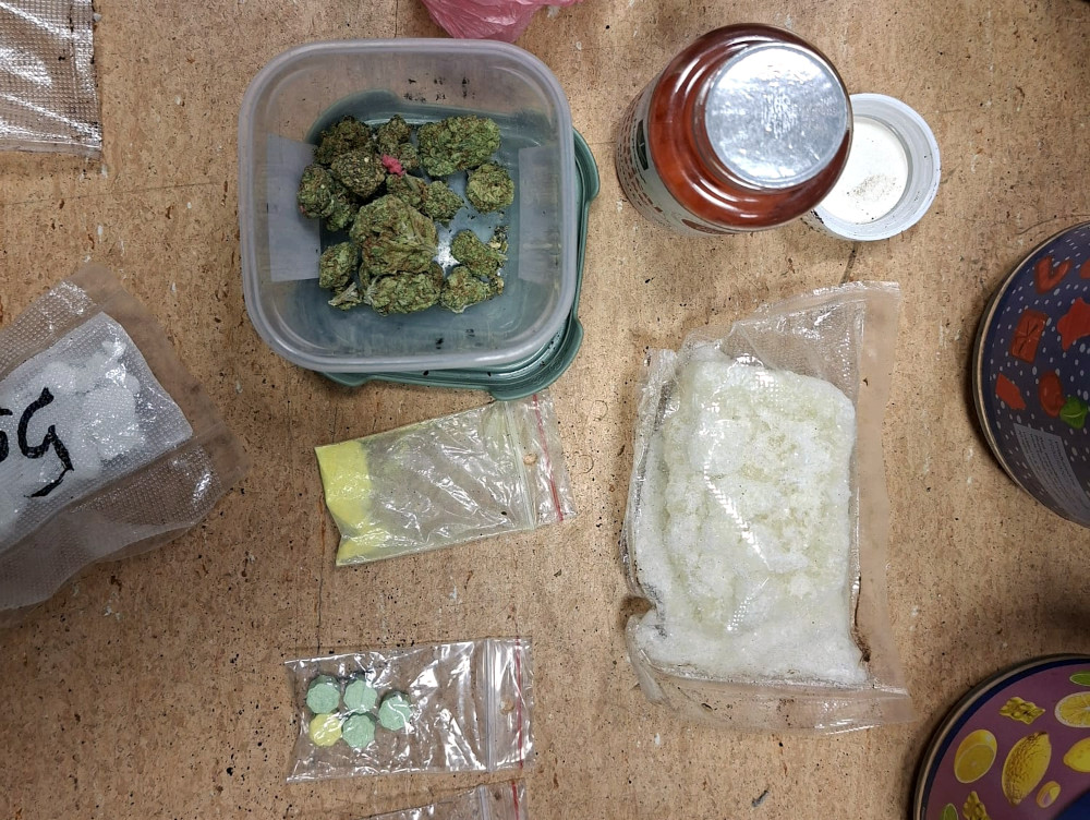 Zielona marihuana w pojemniku obok woreczków z substancjami w proszku i tabletkami na blacie roboczym.