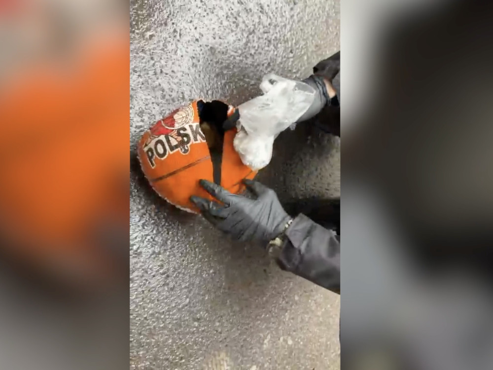 Mężczyzna w rękawiczkach trzyma uszkodzoną piłkę do koszykówki z napisem "POLSKA" na mokrej nawierzchni.