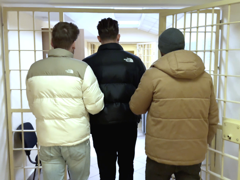 Trzech mężczyzn w kurtkach puchowych przechodzi przez bramkę w budynku, w tle widoczne są kraty.