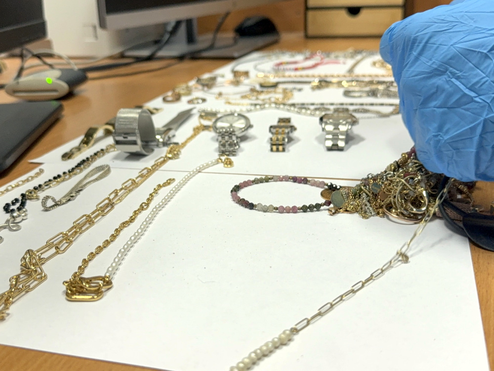 Na białym papierze rozłożone są różnorodne biżuteria i zegarki, a w zbliżeniu widoczna jest ręka w niebieskiej rękawiczce.