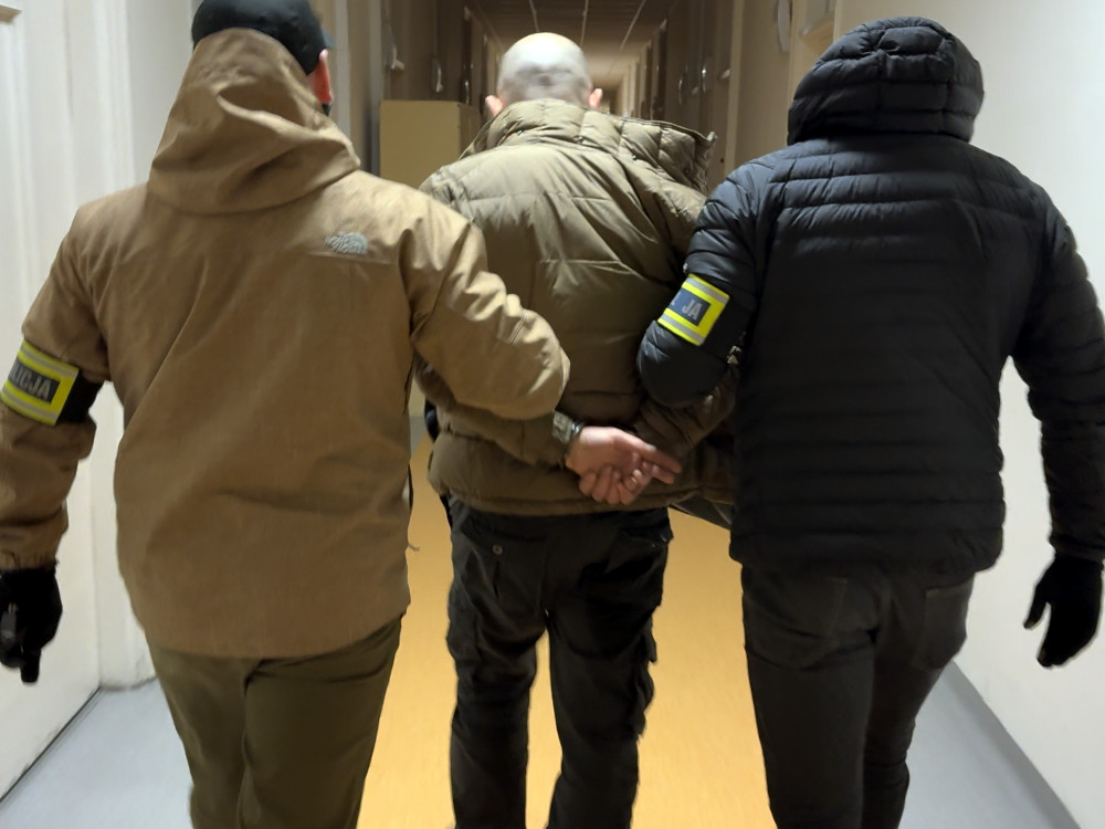 Trzech funkcjonariuszy w mundurach eskortuje aresztowanego mężczyznę korytarzem budynku.