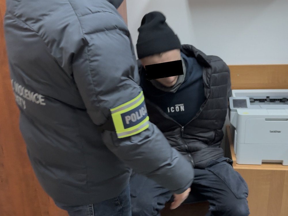 Mężczyzna w czarnej czapce siedzi na krześle, obok niego policjant w szarej kurtce z emblematem "POLICJA".