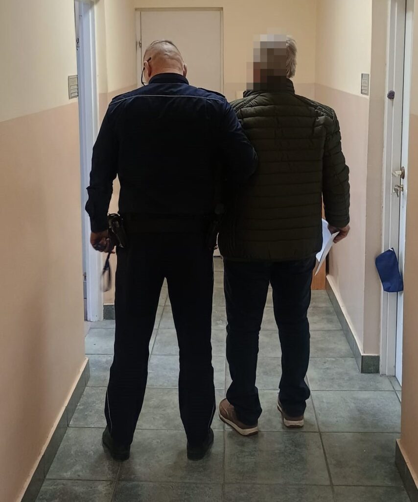 Funkcjonariusz policji prowadzi mężczyznę w zielonej kurtce korytarzem w budynku.