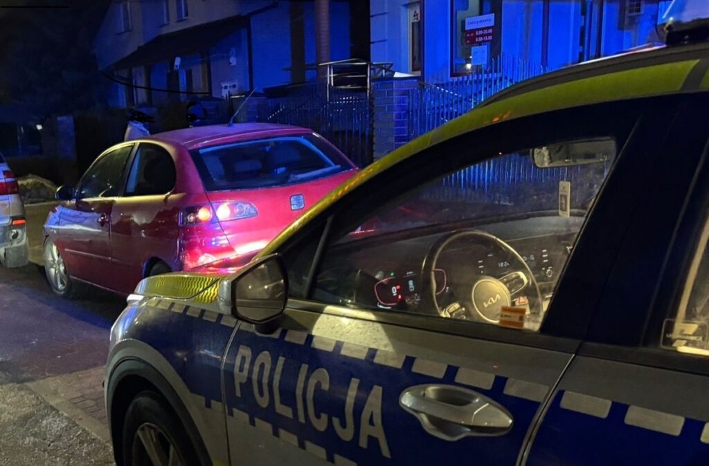 Policyjny samochód z widocznym napisem "POLICJA" zaparkowany obok czerwonego pojazdu na ulicy oświetlonej niebieskimi światłami.