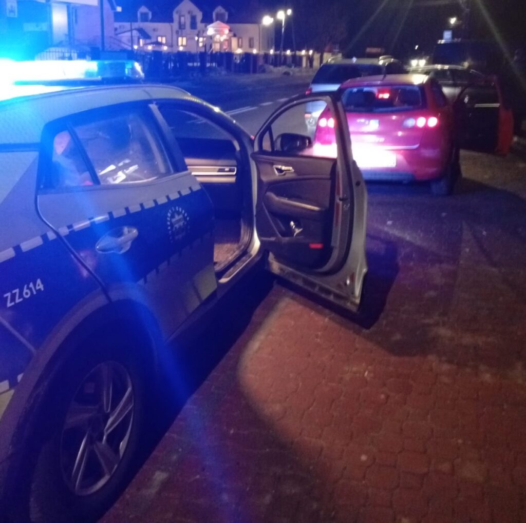 Policyjny radiowóz z włączonymi sygnałami świetlnymi stoi na ulicy nocą, obok zaparkowanego czerwonego samochodu.