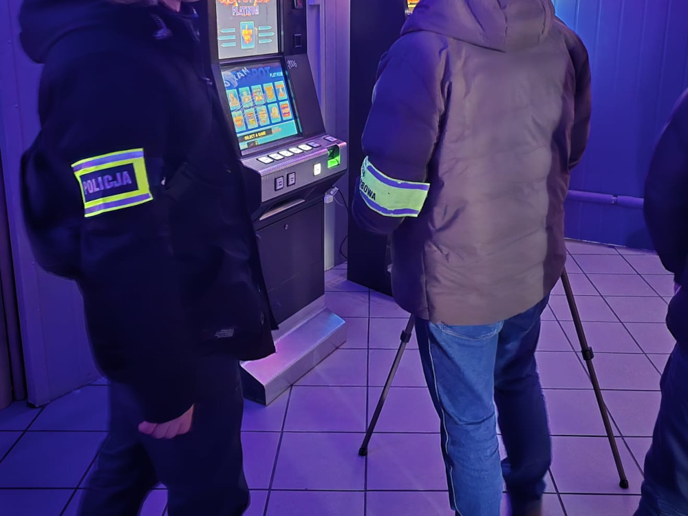 Funkcjonariusze policji w odzieży służbowej obserwują automat do gry w oświetlonym pomieszczeniu.