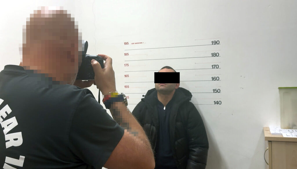 Mężczyzna w czarnej kurtce stoi przy ścianie z oznaczeniami wzrostu, a fotograf robi mu zdjęcie.