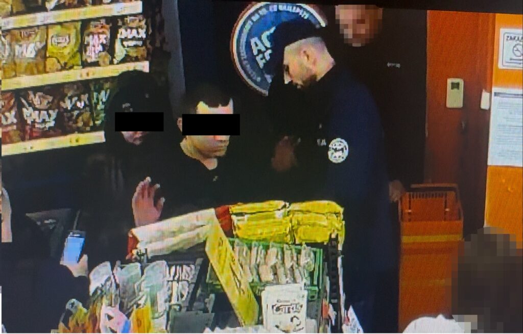 Mężczyzna w czarnej odzieży rozmawia z pracownikiem ochrony w sklepie, w tle półki z przekąskami.