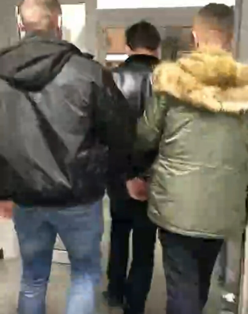 Grupa trzech mężczyzn idzie korytarzem, jeden z nich trzymany jest za rękę przez drugiego.