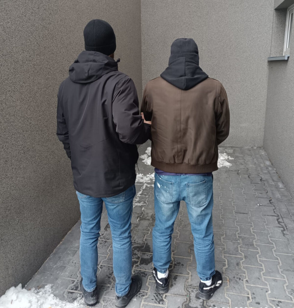 Dwóch mężczyzn w odzieży zimowej stoi plecami do obiektywu w wąskim korytarzu z szarymi ścianami.