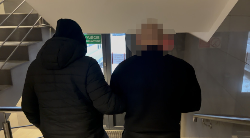 Mężczyzna w czarnej kurtce prowadzi drugiego mężczyznę w kierunku wyjścia z budynku, schody w tle.