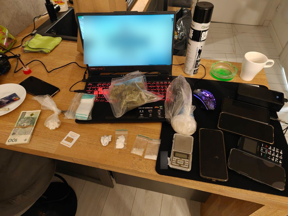 Na stole leżą różne przedmioty, w tym kilka telefonów, woreczki z białym proszkiem, marihuana w przezroczystej torbie, waga oraz banknoty. W tle widoczny jest laptop z włączonym ekranem.