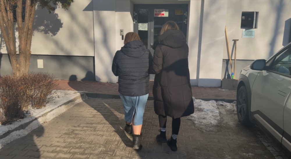 Dwie kobiety idą w stronę wejścia do budynku, jedna w kurtce puchowej, druga w krótkich spodniach.