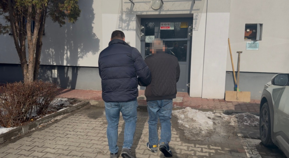 Mężczyzna w kurtce prowadzi drugiego mężczyznę w kierunku wejścia do budynku. W tle widoczny parking z resztkami śniegu.