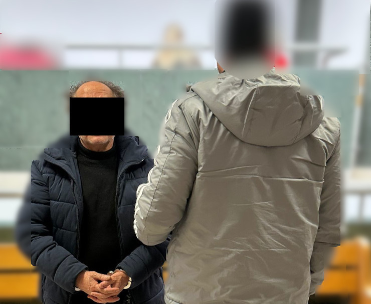 Mężczyzna w ciemnym płaszczu stoi z założonymi rękami, rozmawiając z osobą w szarej kurtce. W tle widoczna jest sala.