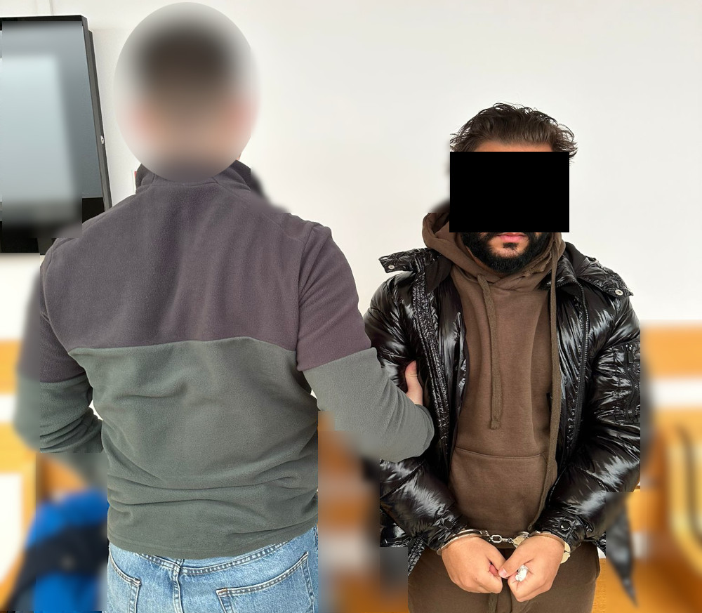 Mężczyzna w kajdankach siedzi na krześle, podczas gdy inny mężczyzna stoi obok niego, trzymając go za ramię.