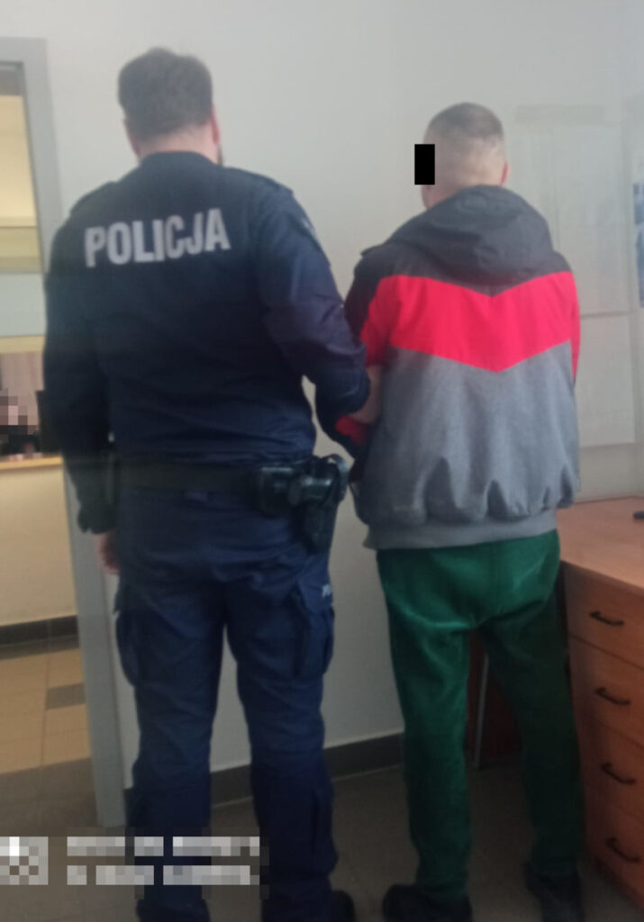Policjant prowadzi zatrzymanego mężczyznę w szarej bluzie z czerwoną wstawką w biurze.