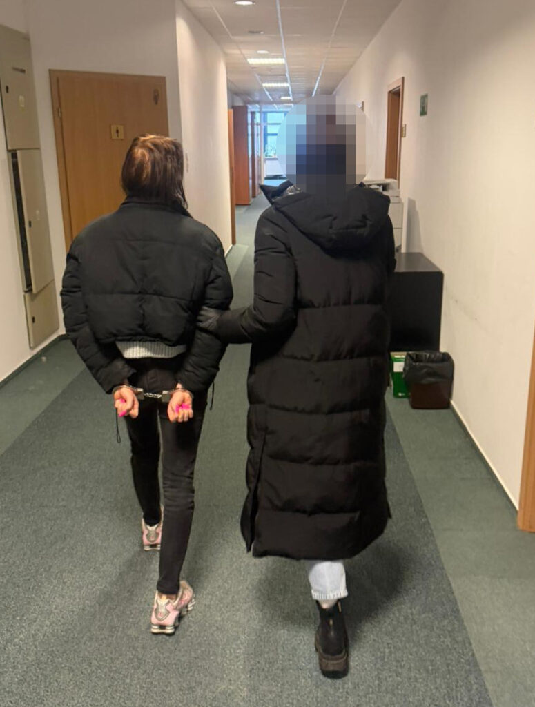 Kobieta w czarnej kurtce prowadzi inną kobietę w kajdankach korytarzem w budynku biurowym.