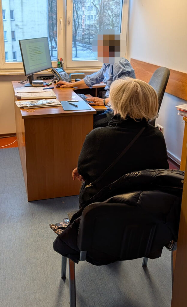 Mężczyzna w niebieskiej koszuli pracuje przy biurku, a kobieta w czarnej kurtce siedzi na krześle, obserwując ekran komputera.