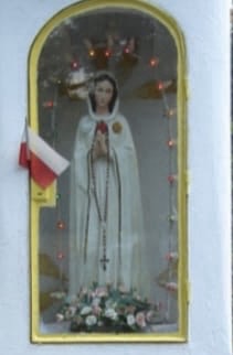 Figura Matki Bożej w białej szacie, otoczona lampkami i kwiatami, umieszczona w niszy na tle białej ściany.