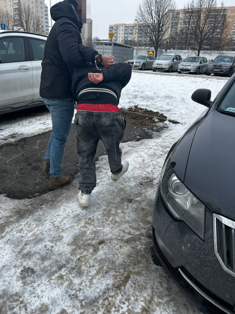 Funkcjonariusz policji zatrzymuje mężczyznę na parkingu w zimowej scenerii, w tle zaparkowane samochody.